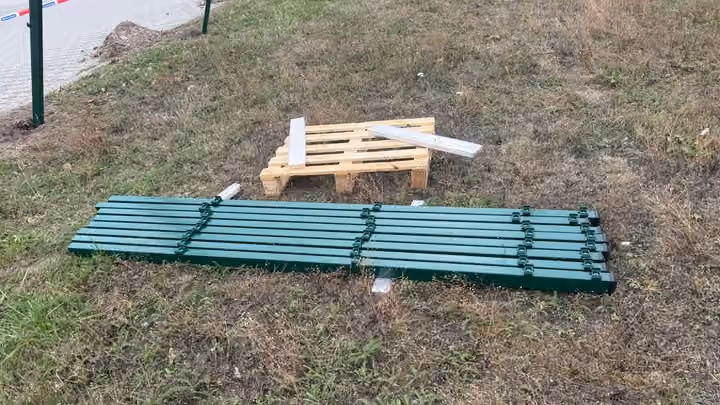 Grün lackierte Einzelteile einer Sitzbank und eine Holzpalette liegen auf einer trockenen Wiese.