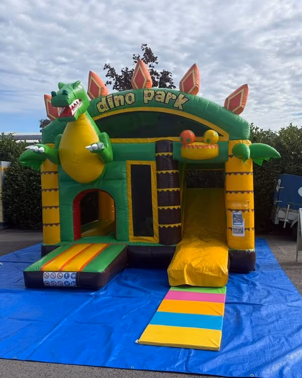 Grün-gelbe Hüpfburg mit einem aufblasbaren Dinosaurier, dem Schriftzug „dino park", einer Rutsche und bunten Matten davor, aufgebaut auf einer blauen Plane vor einer Hecke.