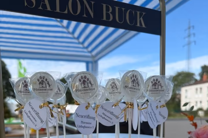 Runde Lutscher mit dem Pfotenwiese-Logo auf einer weißen Etagere, an jedem ein Anhänger „Neueröffnung Pfotenwiese Lüdinghausen e.V."; darüber ein Schild „Salon Buck" unter einem blau-weiß gestreiften Pavillon.