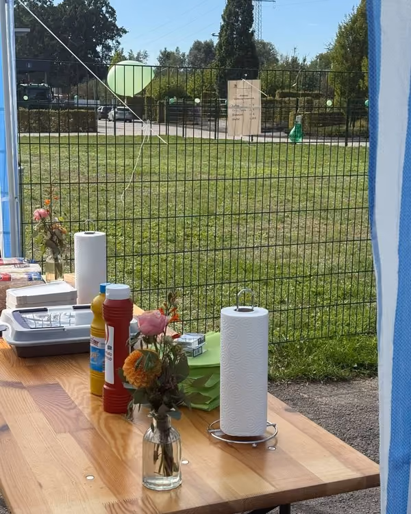 Holztisch mit Blumen in kleinen Glasvasen, Küchenrollen, Senf und Ketchup; dahinter ein grüner Zaun, die Wiese und Luftballons, am Zaun ein Aushang.