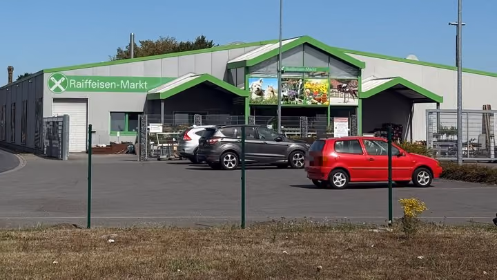 Blick über frisch gesetzte grüne Zaunpfosten hinweg auf den Raiffeisen-Markt mit grünem Schriftzug; davor ein Parkplatz mit geparkten Autos.