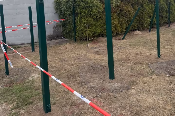 Grüne Zaunpfosten vor einer Betonwand und einer hohen Hecke, davor ein rot-weißes Absperrband und aufgeworfene Erde.