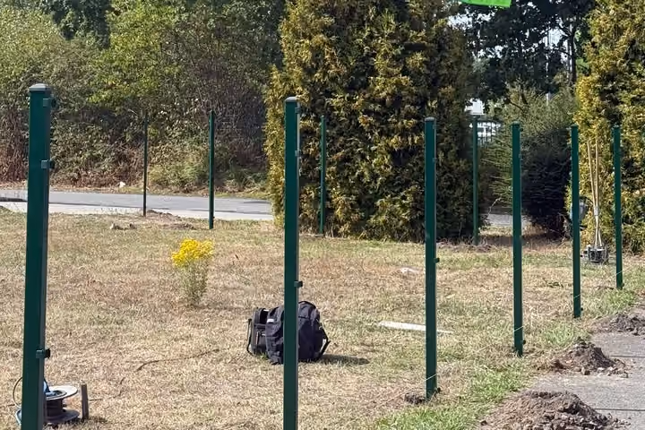 Eine lange Reihe frisch gesetzter grüner Zaunpfosten auf einer abgemähten Wiese; im Gras liegen eine Werkzeugtasche und eine Kabeltrommel.
