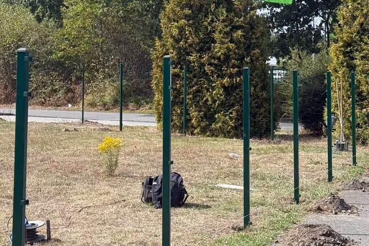 Eine lange Reihe frisch gesetzter grüner Zaunpfosten auf einer abgemähten Wiese; im Gras liegen eine Werkzeugtasche und eine Kabeltrommel.